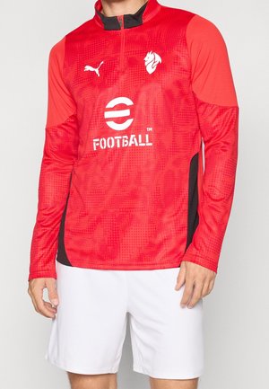 Mand iført rød sportstop med lange ærmer med hvidt fodboldlogo og sorte detaljer, kombineret med simple hvide træningsshorts.