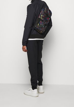 Vue arrière d'une personne portant une veste et un pantalon noirs, avec un sac à dos coloré affichant divers motifs vifs et tourbillonnants.