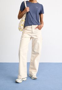 Pantalon larges beige clair associé à un t-shirt bleu marine à manches courtes. Sac texturé avec des accents de fleurs jaunes. Baskets grises et blanches.