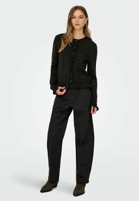 Cardigan noir en mailles avec un col rond, un devant froncé et une fermeture à boutons. Associé à un pantalon noir ample et des bottines à talons pointus.