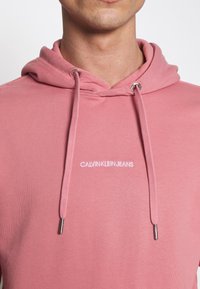 Felpa con cappuccio rosa con coulisse, occhielli argentati e logo bianco "CALVIN KLEIN JEANS" ricamato sul petto. Texture morbida.