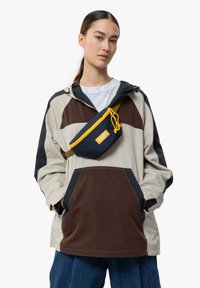 Giacca leggera con blocchi di colore in beige, marrone e blu navy, dotata di cappuccio e grandi tasche. Abbinata a una borsa a fanny pack blu navy con dettagli gialli.