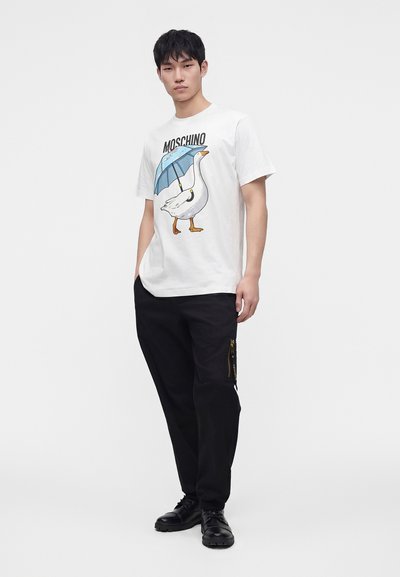 Νέος άνδρας που φοράει λευκό t-shirt Moschino με εκτύπωση μιας χήνας που κρατάει μια μπλε ομπρέλα, μαύρο παντελόνι με τσέπη με φερμουάρ και μαύρα παπούτσια.
