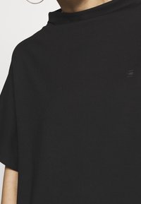 Lyhythihainen musta paita, joka on valmistettu sileästä kankaasta. Siinä on korkea pääntie ja pieni logo etupuolella. Minimalistinen muotoilu.