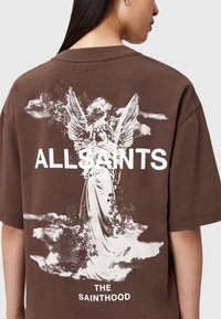 Camiseta de algodón marrón con un gráfico blanco de un ángel con alas y nubes. El texto incluye "ALLSAINTS LONDON" y "THE SAINTHOOD."