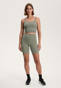 Olivgrön sport-bh och högmidjade shorts-set, med en åtsittande design, slät yta och justerbara remmar. Svarta träningsskor på.