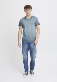 Camiseta gris de cuello en V con un acabado texturizado, combinada con jeans azul claro y zapatillas de deporte en blanco y negro. De pie contra un fondo liso.