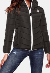 Zwarte pufferjack met capuchon, voorzien van een witte ritssluiting, diagonale gestikte patroon en zijzakken. G gedragen over blauwe distressed jeans.