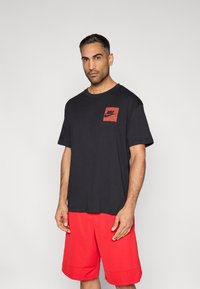 Μαύρο βαμβακερό μπλουζάκι με κόκκινο τετράγωνο λογότυπο στο στήθος, συνδυασμένο με κόκκινα σορτς. Χαλαρή γραμμή, λαιμόκοψη τύπου crew και κοντά μανίκια.