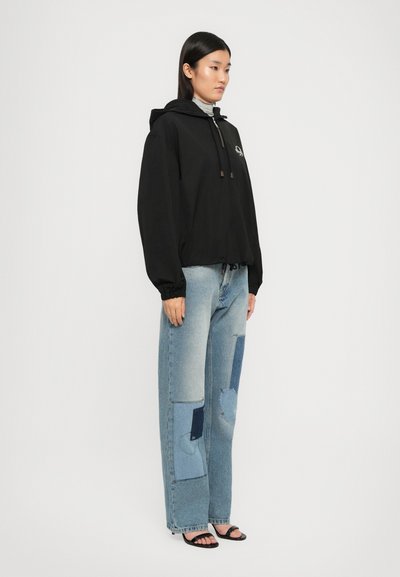 Črna jakna s kapuco in vezalkami, v kombinaciji s svetlo modrimi jeans hlačami s različnimi denim zakladi. Model nosi črne sandale.