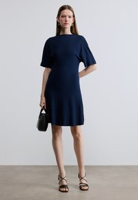 Rochie bleumarin cu mâneci scurte, material neted, lungime până la genunchi, cu guler rotund. Modelul ține o geantă mică neagră și poartă sandale negre cu bretele.