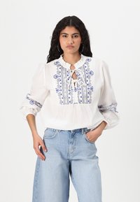 Blouse blanche avec broderies florales bleues, encolure froncée et détail en trou de serrure. Manches longues avec poignets élastiques. Portée avec un jean bleu clair.