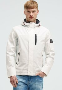 Giacca impermeabile bianca con cappuccio, dettagli con cerniera nera e tasche laterali con bottone. Presenta una patch con il logo sulla manica.