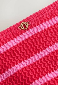 Rood getextureerde stof met geplooide strepen in verschillende tinten roze, afgewerkt met een goudkleurige ronde sluiting.