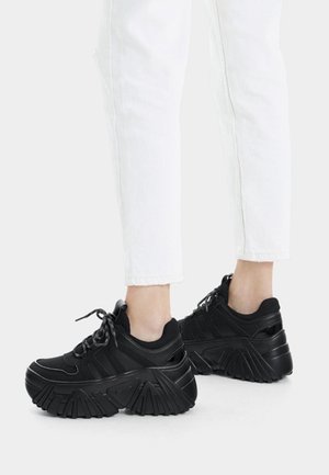 Stopy w czarnych masywnych sneakerach na platformie z grubymi podeszwami, w połączeniu z białymi, krótkimi spodniami na jednolitym tle.