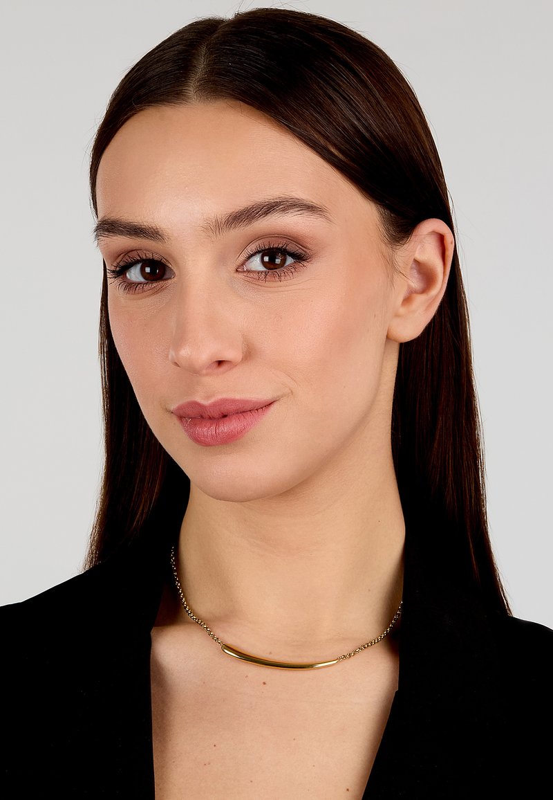 Collana in metallo dorato con pendente a barra curva su una catena, che riposa sulla clavicola, con una finitura liscia e un leggero scintillio.