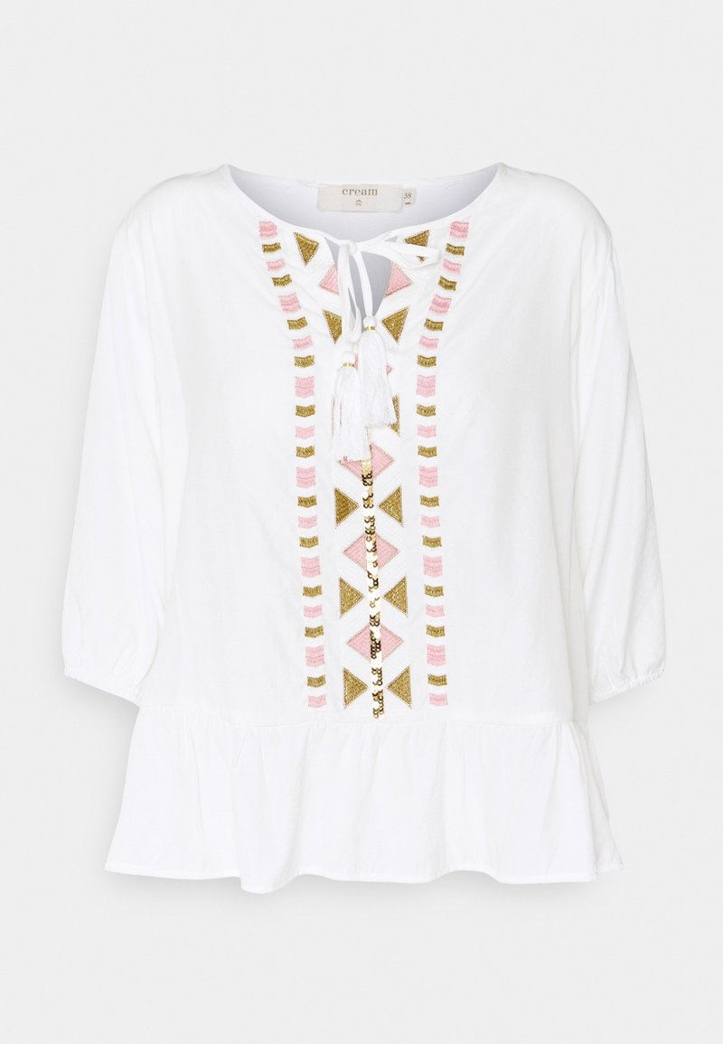 Blouse blanche à manches trois-quarts, ourlet à volants, encolure à nouer devant et broderie géométrique rose, dorée et beige au centre.