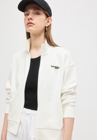 Νεαρή γυναίκα με λευκό μπουφάν Lacoste με φερμουάρ, μαύρο πουκάμισο, λευκό παντελόνι, μαύρο καπέλο και χρυσούς κρίκους σκουλαρίκια, μπροστά σε απλό ανοιχτό φόντο.