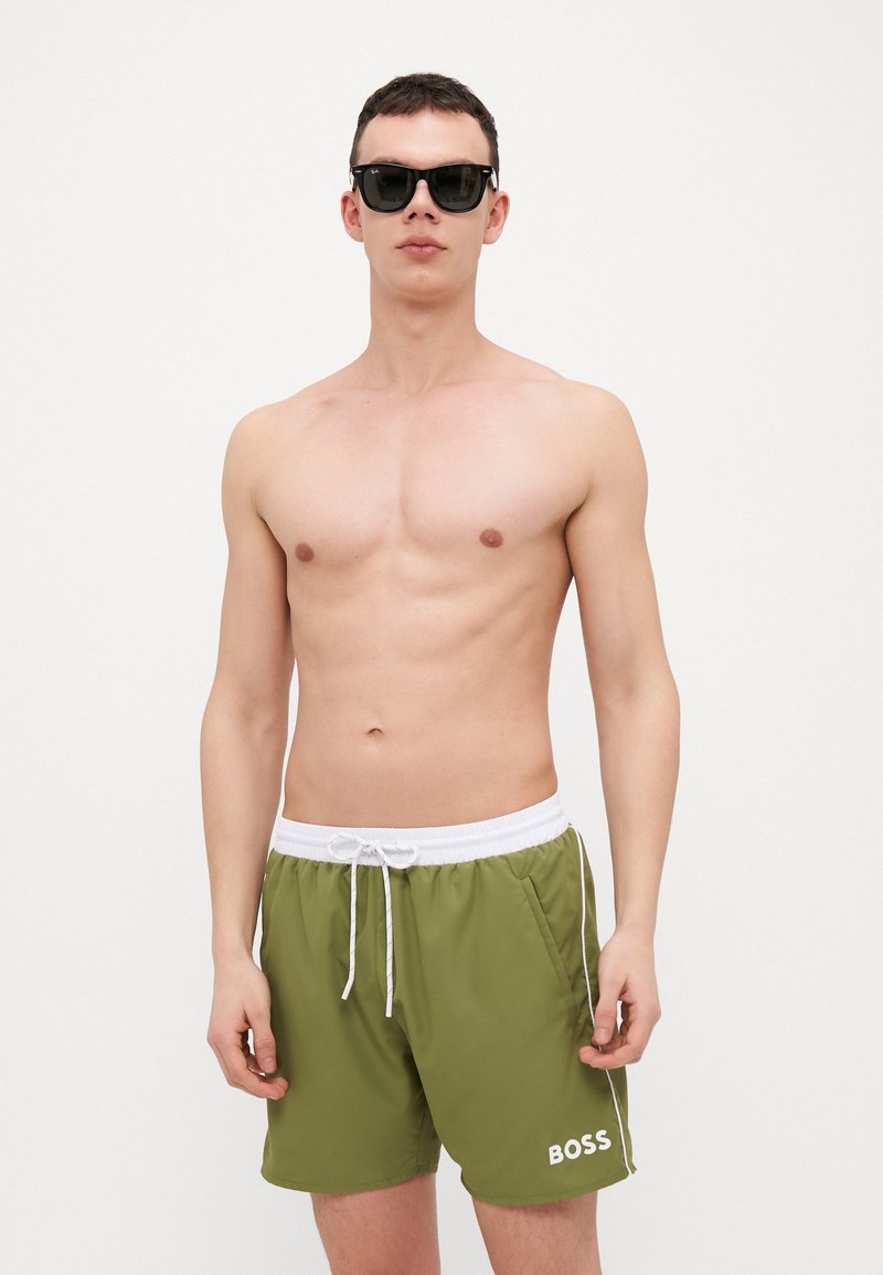 Costume da bagno verde con laccetti bianchi e dettagli laterali, con logo "BOSS". Il modello indossa occhiali da sole neri su uno sfondo neutro.