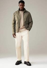 Jachetă bomber verde măsliniu cu guler înalt, pulover bej dedesubt, pantaloni albi și pantofi cu talpă groasă negri. Design simplu și funcțional.