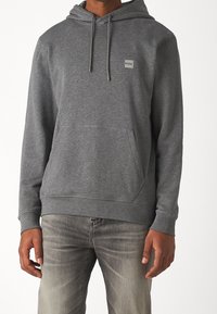 Grey hoodie made of soft fabric with a front pocket, drawstring hood, and a small brand logo patch on the chest. Worn with light jeans.