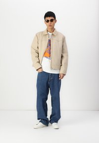 Chaqueta beige con cremallera y cuello, llevada sobre una camiseta blanca con un gráfico en naranja y azul, combinada con jeans azules anchos y zapatillas blancas.