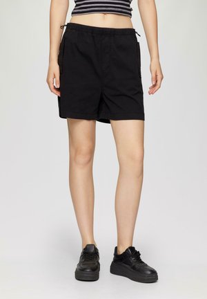 Shorts noirs avec une taille élastique et un cordon de serrage. Comprend des poches latérales et une coupe ample. Associés à des baskets noires.