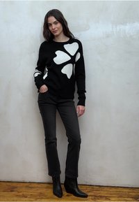 Zwarte trui met groot wit bloemmotief, gecombineerd met zwarte jeans en enkellaarsjes. De textuur van de trui is zacht en gebreid.