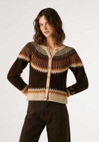 Cardigan marron avec un motif géométrique dans des tons d'orange et de beige. Présente un col rond, une fermeture à bouton et des poignets côtelés.