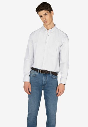 Camisa de cuello blanco con rayas verticales azules, combinada con unos vaqueros de un azul claro y un cinturón oscuro trenzado. La camisa presenta un pequeño logo en el pecho.