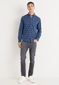 Camicia a quadri blu con colletto, jeans grigi slim-fit e sneakers bianche. La camicia presenta una tasca e una texture di tessuto morbido.