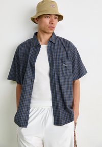 Chemise à manches courtes, boutonnée, à motif vichy bleu marine et bleu clair, avec une poche poitrine arborant un logo. Portée par-dessus un débardeur blanc.