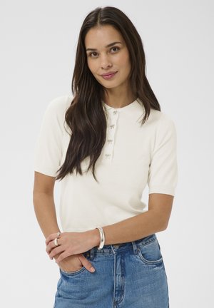 Jeune femme aux longs cheveux noirs portant un polo blanc à manches courtes et un jean bleu, souriante avec les mains doucement jointes devant elle.