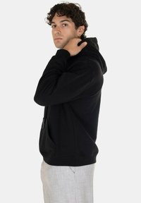 Felpa nera con cappuccio e coulisse, tasca frontale, polsini e orlo a costine. Indossata con pantaloni grigi chiari; il modello posa di profilo su uno sfondo bianco.