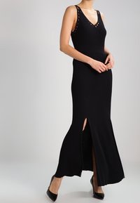 Abito lungo nero in maglia con scollatura a V profonda, spalline testurizzate e spacco laterale. Indossato con tacchi neri, evidenziando la silhouette aderente e il tessuto morbido.