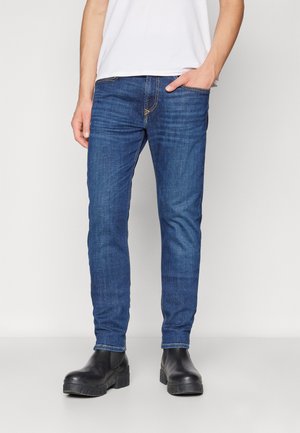 Mand iført blå slim fit jeans med synlige syninger, sorte chunky støvler og en hvid skjorte, stående foran en ensfarvet hvid baggrund.