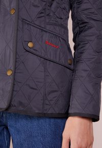 Prošívaná námořnická bunda s texturovaným povrchem, obsahující náprsní kapsu, bronzové patentky a červené vyšívané logo. Lemována černým okrajem.