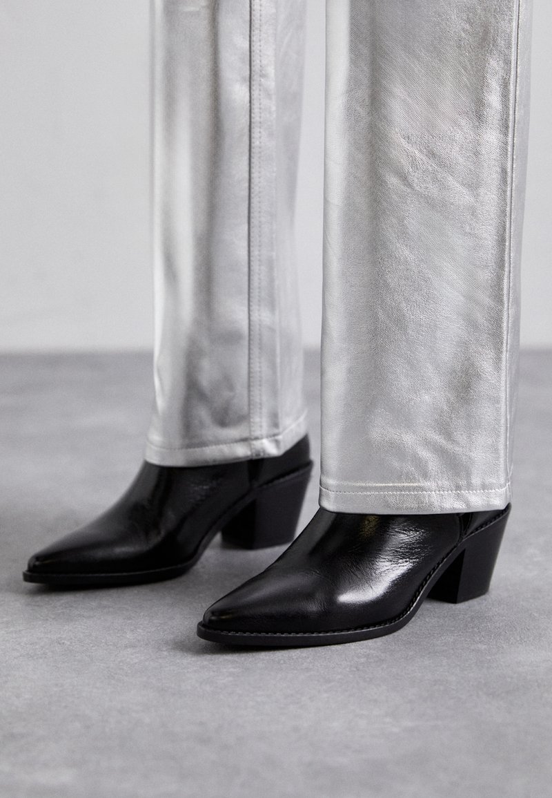 Bottes noires en cuir à talon carré et bout pointu, associées à un pantalon argenté brillant, mettant en valeur des textures épurées et des couleurs contrastées.