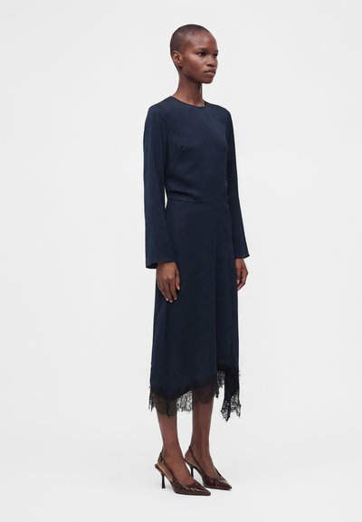 Femme debout portant une robe bleu marine à manches longues avec un ourlet en dentelle asymétrique et des talons pointus marron sur fond uni.