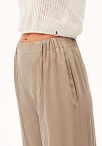 Volné béžové kalhoty s elastickým pasem, bočními kapsami a plisovaným designem. Tkanina má hladký povrch; malý logo detail na kapse.