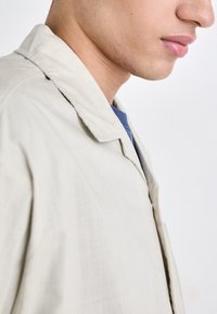 Chaqueta beige de tela suave, diseño con cuello y detalle de botones en la parte frontal, llevada sobre una blusa azul, mostrando una silueta relajada.