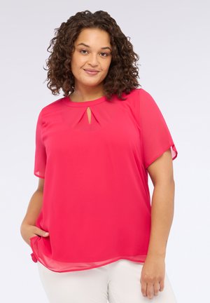 Frau mit lockigem Haar, die eine leuchtend pinke Kurzarmbluse mit einem Schlüssel-Design-Ausschnitt trägt, steht mit einer Hand in der Tasche und lächelt.