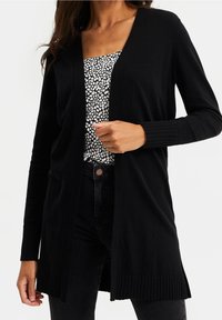 Svart stickad cardigan med sjalkrage, ribbade ärmslut och nederkant. Öppen framsida med avslappnad passform, lagd över en mönstrad topp.