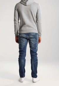 Grå huvtröja, blå denimjeans med en urtvättad look, bakvy som visar två bakfickor och vita sneakers.