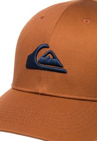 Brun baseballkeps med en texturerad, ribbad yta. Har en marinblå broderad logotyp framtill. Skärmen och ventilationshål är synliga.
