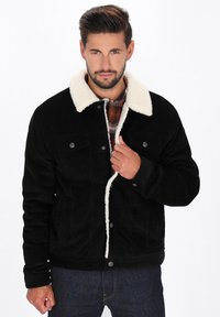 Schwarze Cordjacke mit einem weißen Lammfellkragen, vorderer Knopfverschluss, Brusttaschen und strukturiertem Stoff, getragen über einem karierten Hemd.