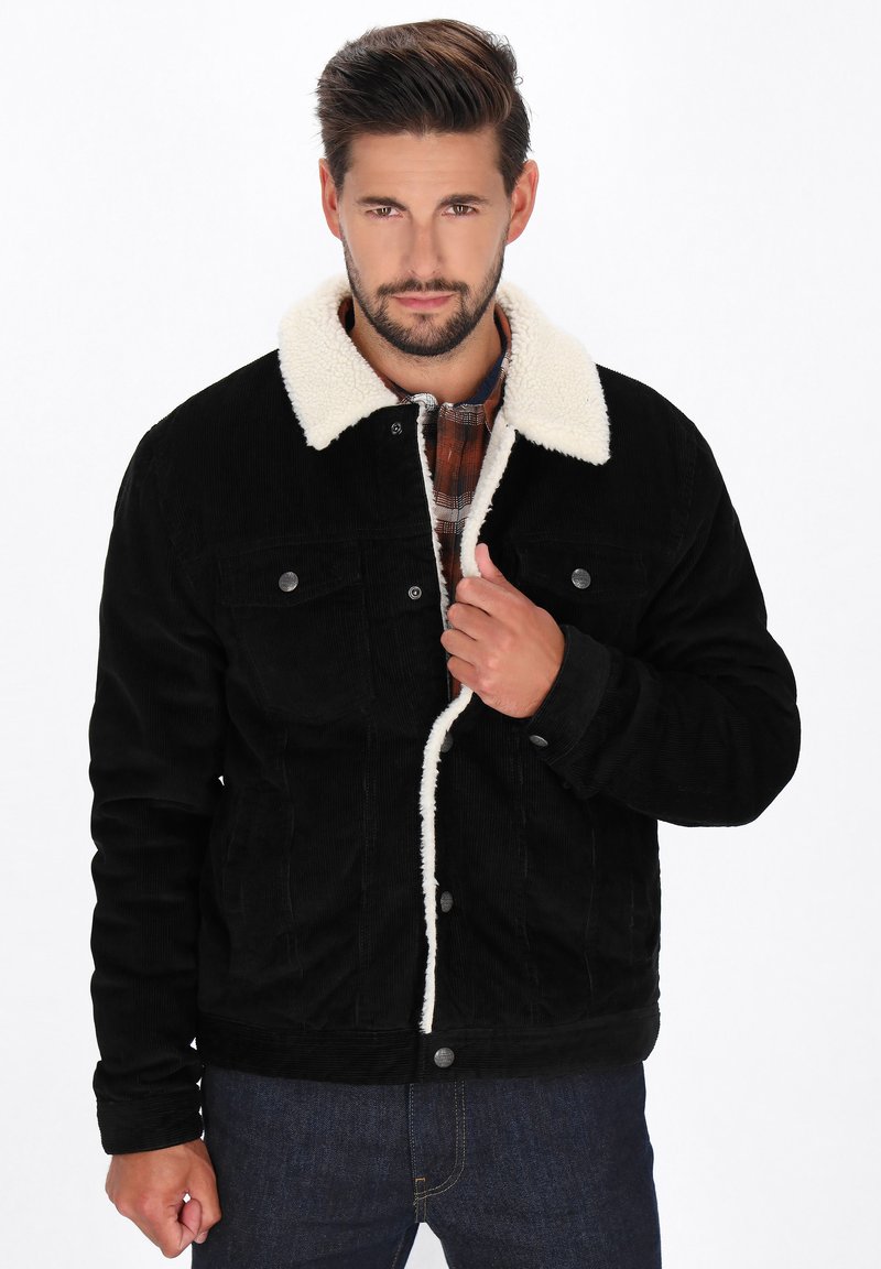 Schwarze Cordjacke mit einem weißen Lammfellkragen, vorderer Knopfverschluss, Brusttaschen und strukturiertem Stoff, getragen über einem karierten Hemd.