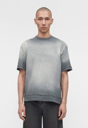 Άνδρας με γκρι μπλουζάκι με σύντομο μανίκι σε βαθμίδα και σκούρα γκρι παντελόνια, στέκεται μπροστά σε ένα απλό φωτεινό φόντο.