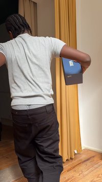 Personne aux cheveux tressés portant une chemise grise et un pantalon noir, tenant une boîte à chaussures Vans bleue dans une pièce avec un sol en bois et des rideaux jaunes.