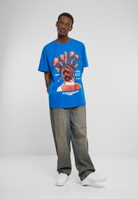 Blå grafisk t-shirt med rød og sort print, vist på en model iført lysegrå baggy jeans og hvide sneakers.